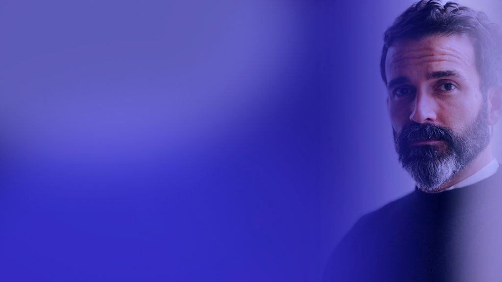 Hombre con barba mirando a cámara sobre un fondo degradado azul violeta
